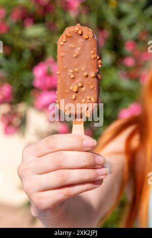 Eine Nahaufnahme einer Hand, die ein Schokoladeneis in der Hand hält, hervorgehoben vor einem lebhaften Hintergrund aus rosa Blumen, die die Freude symbolisieren Stockfoto