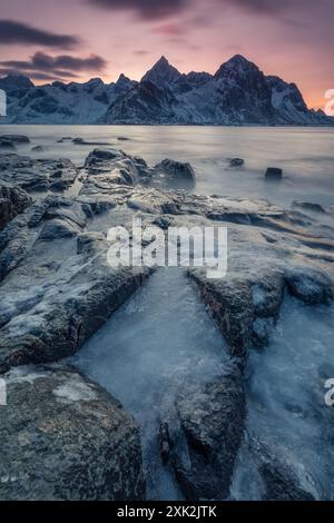 Eine ruhige, winterliche Szene fängt das sanfte Sonnenaufgangslicht vor eisigen Felsen und majestätischen Bergen in Lofoten, Norwegen, ein und hebt die raue Natur der Region hervor Stockfoto