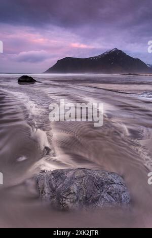 Ein atemberaubender Blick auf einen ruhigen Strand in Lofoten unter einem Dämmerungshimmel. Das Bild zeigt dynamische Wasserpfade, die zu einem einsamen Felsen mit einem Drama führen Stockfoto