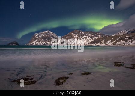 Ein atemberaubender Blick auf die Nordlichter, die sich über weiße, schneebedeckte Berge neben einem ruhigen Strand in Lofoten, Norwegen, erheben Stockfoto