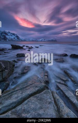 Ätherischer Sonnenuntergang über einem felsigen Strand in Lofoten, Norwegen, mit weichen rosa Wolken, schneebedeckten Bergen und einem nebeligen Meer, das durch Langzeitfotos erzeugt wurde Stockfoto