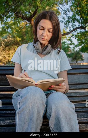 Eine junge Frau schreibt in einem Notizbuch und sitzt an einem sonnigen Tag auf einer Parkbank mit ihren Kopfhörern um den Hals. Stockfoto