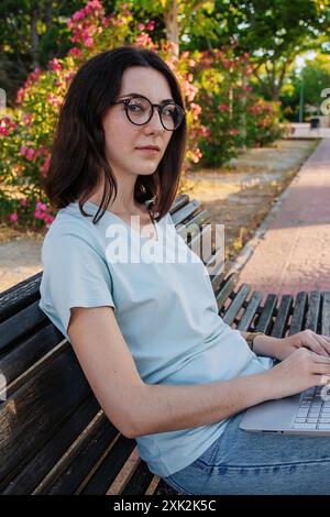 Seitenansicht einer jungen Frau mit dunklem Haar und Brille mit einem Laptop, während sie auf einer Parkbank sitzt, umgeben von rosa Blumen und Grün. Stockfoto