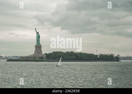 Die legendäre Freiheitsstatue steht hoch auf Liberty Island, mit einem kleinen Segelboot im Vordergrund, der bewölkte Himmel und das ruhige Wasser schaffen einen Stockfoto