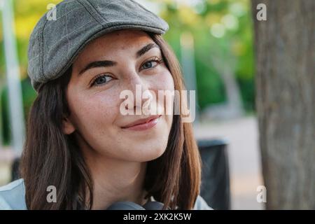 Nahaufnahme einer jungen Frau, die eine graue Mütze oder Baskenmütze trägt und lächelt und direkt auf die Kamera im Freien blickt. Stockfoto