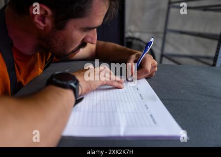 Ein fokussierter Mann, der ein lässiges oranges Hemd und eine Sportuhr trägt, wird in einer dunklen, büroähnlichen Umgebung erfasst, indem er Dokumente oder Arbeitsblätter ausfüllt Stockfoto