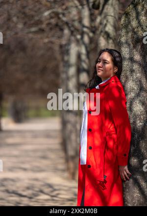 Porträt einer Frau draußen. Eine kaukasische Dame in rotem Mantel und weißem Hemd steht an einem Baum und schaut nachdenklich in die Ferne. Stockfoto