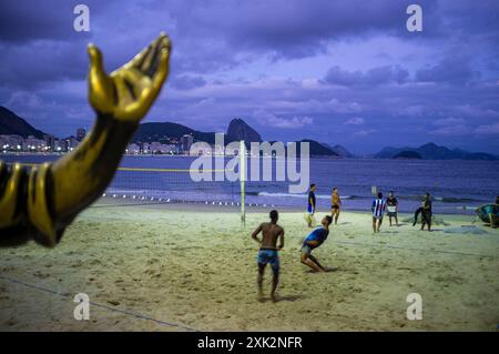 Strandkultur am Strand Copacabana - Jungen spielen mitten im Geschehen Footvolley - Beachvolleyball-Spiel, das nur mit Füßen, Brust und Kopf gespielt wird, um den Ball zu schlagen - während eine andere Gruppe von Jungen direkt hinter der ersten Gruppe Keep-ups oder Kick-ups spielt; die Fähigkeit, mit einem Fußball zu jonglieren, indem man Füße, Unterschenkel, Knie, Brust, Schultern und Kopf benutzt, ohne dass der Ball auf den Boden trifft. Der Arm der Dorival Caymmi Statue im Vordergrund an der Copacabana Strandpromenade und dem Zuckerhut im Hintergrund, Rio de Janeiro, Brasilien. Stockfoto