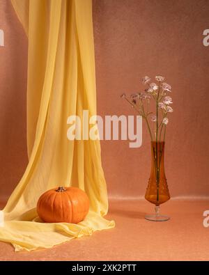 Ruhiges Stillleben. Ein Blumenzweig in einem transparenten braunen Glas, eine Vase, ein Kürbis daneben, ein gelber Vorhang hängt auf einem orangefarbenen Hintergrund. Stockfoto