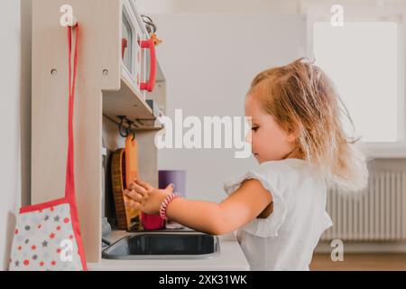 Das junge Mädchen spielt glücklich in einem gemütlichen Spielzimmer mit ihrer bunten Spielzeugküche. Mit ihrer Fantasie kochen und Geschirr waschen. Ganz in ihren Preten eingetaucht Stockfoto