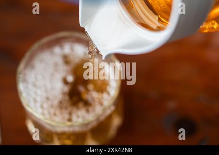 Eine Nahaufnahme eines Glases, das mit einem goldenen Getränk aus einem weißen Krug gefüllt wird. Das Getränk ist sprudelnd und wirkt erfrischend, mit einer hölzernen Brandung Stockfoto