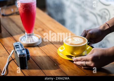 Eine Person, die eine gelbe Tasse Latte Art auf einem Holztisch neben einem rosafarbenen Getränk in einem hohen Glas platziert. Eine Vintage-Kamera steht ebenfalls auf dem Tisch und schafft Stockfoto