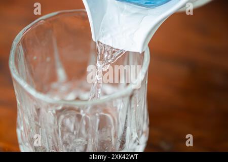 Ein klares Glas, das mit Wasser aus einem Pitcher gefüllt wird. Das Wasser strömt hinein und erzeugt Wellen im Glas. Der Hintergrund ist aus Holz und sorgt für eine warme Atmosphäre Stockfoto