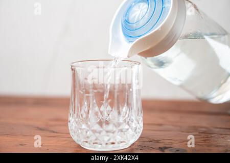 Ein klares Glas, das mit Wasser aus einem Pitcher gefüllt wird. Der Pitcher hat einen blauen Deckel und gießt Wasser in ein strukturiertes Glas auf einer Holzoberfläche. Stockfoto