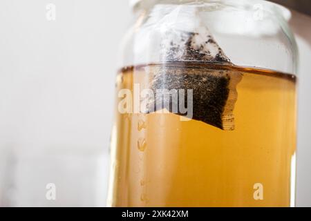 Eine Nahaufnahme eines Glasbehälters mit gebrühtem Tee, mit einem Teebeutel, der in die Flüssigkeit getaucht ist. Der Tee hat eine kräftige bernsteinfarbene Farbe und das Glas ist teilweise gefiltert Stockfoto