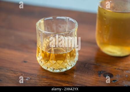 Eine Nahaufnahme eines mit bernsteinfarbener Flüssigkeit gefüllten Glases auf einem Holztisch. Ein Gefäß mit der gleichen Flüssigkeit ist teilweise im Backgro sichtbar Stockfoto