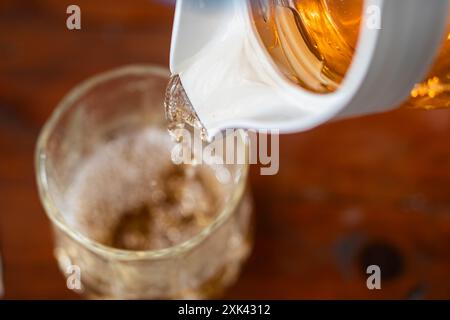 Eine Nahaufnahme eines Glases, das mit einer goldbraunen Flüssigkeit aus einem weißen Pitcher gefüllt wird. Die Flüssigkeit scheint ein erfrischendes Getränk zu sein, möglicherweise geeist Stockfoto