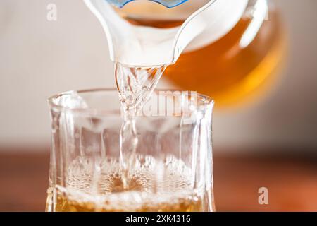 Eine Nahaufnahme eines klaren Glases, das mit einer goldenen Flüssigkeit aus einem Pitcher gefüllt wird. Die Flüssigkeit ist sprudelnd und brausend, was darauf hindeutet, dass es sich um ein Kohlendioxid handeln könnte Stockfoto