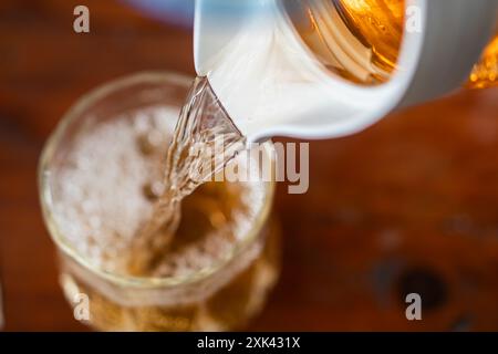 Eine Nahaufnahme eines Getränks, das aus einem weißen Krug in ein Glas gegossen wird. Das Getränk scheint eine goldbraune Farbe mit Blasen zu sein, was darauf hindeutet Stockfoto