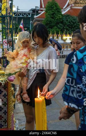 Bangkok, Thailand. Juli 2024. Eine Mutter mit ihrer Tochter in den Armen, die in einem Regenmantel gekleidet ist, wird während der Zeremonie des Asalha Puja Day im Wat Thepleela Tempel gesehen, wie sie ihre Kerze zündet. Asalha Puja, auch bekannt als Dharma Day, ist eines der wichtigsten Feste des Theravada-Buddhismus und feiert Buddhas erste Predigt. Der Tag wird als Gelegenheit für Buddhisten gesehen, ihre Dankbarkeit zu zeigen, indem sie Tempelopfer spenden und Predigten lauschen. Quelle: SOPA Images Limited/Alamy Live News Stockfoto