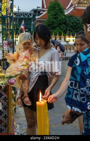 Bangkok, Thailand. Juli 2024. Eine Mutter mit ihrer Tochter in den Armen, die in einem Regenmantel gekleidet ist, wird während der Zeremonie des Asalha Puja Day im Wat Thepleela Tempel gesehen, wie sie ihre Kerze zündet. Asalha Puja, auch bekannt als Dharma Day, ist eines der wichtigsten Feste des Theravada-Buddhismus und feiert Buddhas erste Predigt. Der Tag wird als Gelegenheit für Buddhisten gesehen, ihre Dankbarkeit zu zeigen, indem sie Tempelopfer spenden und Predigten lauschen. (Foto: Nathalie Jamois/SOPA Images/SIPA USA) Credit: SIPA USA/Alamy Live News Stockfoto