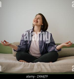 Glückliche, große Frau, die in meditativer Pose auf dem Bett sitzt. Sie lächelt mit geschlossenen Augen, heiterer und fröhlicher Atmosphäre. Themen der Meditation, Selbstpflege und psychischen Wohlbefinden. Stockfoto