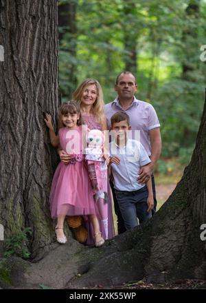 Porträt einer glücklichen Familie. Ein lachender Vater und eine Mutter, ein Teenager-Sohn und eine Erstklässler-Tochter stehen neben einem großen Baum im Park. Mutter und Mädchen Stockfoto