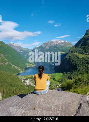 Eine asiatische Frau sitzt auf einem felsigen Felsvorsprung mit Blick auf einen atemberaubenden norwegischen Fjord, umgeben von üppigen grünen Bergen und einem klaren blauen Himmel. Geiranger Fjord Norwegen Stockfoto