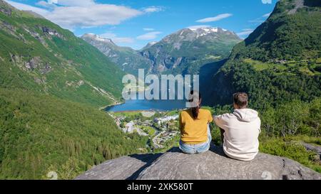 Zwei Personen sitzen auf einem felsigen Felsvorsprung mit Blick auf einen atemberaubenden norwegischen Fjord, umgeben von üppigen grünen Bergen und einem klaren blauen Himmel. Geiranger Fjord Norwegen Stockfoto