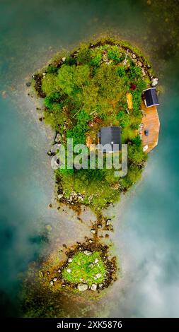 Ein Blick aus der Vogelperspektive auf eine kleine, grüne Insel in Norwegen, mit einer Hütte und einer Holzterrasse mit Blick auf das nebelgefüllte Wasser. Lovrafjorden, Norwegen Stockfoto