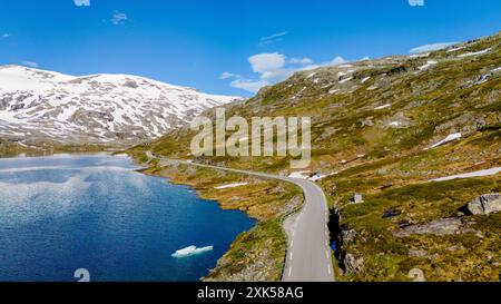 Eine malerische Straße schlängelt sich durch eine bergige Landschaft in Norwegen. Die Straße umgibt den Rand eines glitzernden blauen Sees mit schneebedeckten Bergen im Hintergrund. Strynefjellsvegen, Geiranger, Norwegen Stockfoto