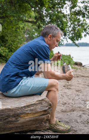 Ein Mann mittleren Alters reist, sitzt am Ufer des Sees, isst Nahrung aus einer Thermoskanne. Meditation, Genuss, Entspannung. Stockfoto