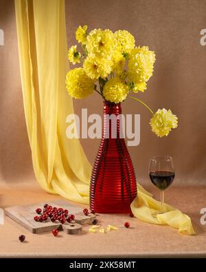 Ein großer Strauß gelber Blumen in einer roten Vase, neben einem Glas Rotwein und roten Beeren auf orangefarbenem Hintergrund und einem hängenden gelben Schal Stockfoto