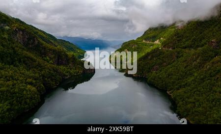 Ein dramatischer Blick auf einen ruhigen Fjord, eingebettet in majestätische grüne Berge. Wolken ziehen über sich, ihre Schatten fallen auf das ruhige Wasser. Lovrafjorden, Norwegen Stockfoto