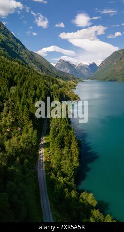 Eine gewundene Straße schlängelt sich durch einen üppigen Wald, der an einen lebhaften türkisfarbenen Fjord grenzt. Schneebedeckte Berge erheben sich in der Ferne und schaffen eine atemberaubende Landschaft. Fjaerlandsfjorden, Vestland, Norwegen Stockfoto