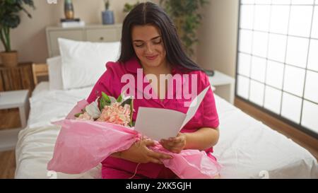 Frau im rosa Pyjama, die auf dem Bett im Schlafzimmer sitzt, eine Lesekarte hat und einen Blumenstrauß hält. Stockfoto