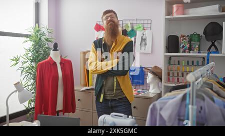 Ein selbstbewusster roter Mann mit Bart steht in einem bunten Schneidergeschäft, umgeben von Kleidungsstücken und Nähzubehör, die Arme überkreuzt. Stockfoto
