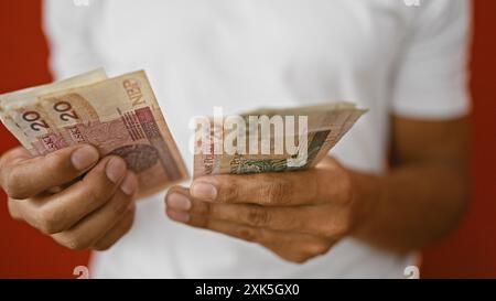 Junger hispanischer Mann, der polnische Zloty-Banknoten an einer roten Wand hält und ein Gefühl von Handel und Transaktion vermittelt. Stockfoto