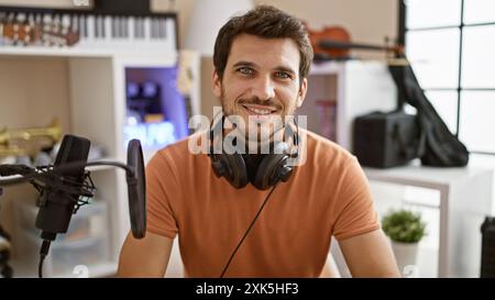 Ein lächelnder junger Mann mit Kopfhörern um den Hals posiert in einem gut ausgestatteten Musikstudio. Stockfoto