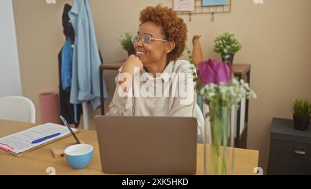 Eine attraktive afroamerikanische Frau, die von zu Hause aus arbeitet, lächelt und in Gedanken an ihrem überfüllten Schreibtisch verlor. Stockfoto