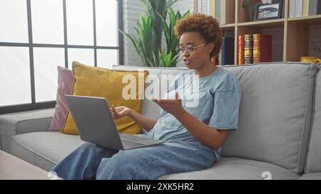 Eine attraktive schwarze Frau in lässiger Kleidung führt zu Hause einen Videoanruf und sitzt bequem auf einer Couch mit hellen Kissen und einem Laptop. Stockfoto