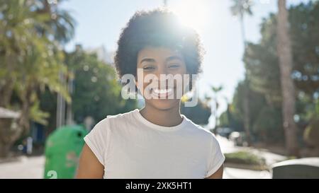 Eine fröhliche afroamerikanische Frau mit lockigen Haaren, die sich in einer sonnigen Umgebung im Freien posiert und Selbstvertrauen und Glück ausstrahlt. Stockfoto