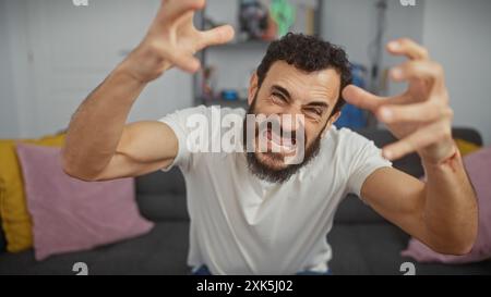 Bärtiger Mann mittleren Alters in weißem T-Shirt mit aggressiver Geste im modernen Wohnzimmer Stockfoto