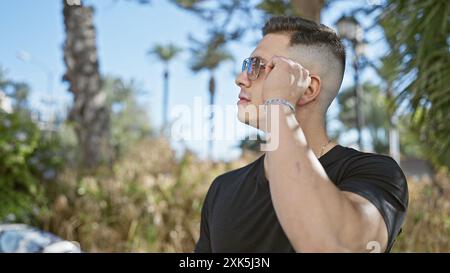 Ein junger hispanischer Mann passt seine Sonnenbrille in einem sonnigen Park mit Palmen an. Stockfoto