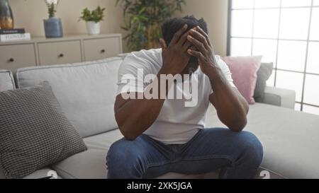 Ein junger afroamerikaner mit Bart sitzt in einem Wohnzimmer, hält den Kopf in den Händen, erscheint verstört, in einer modernen, dekorierten Wohnung Stockfoto