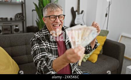 Ein lachender Mann mittleren Alters, der britische Pfund hält, sitzt drinnen in einem Wohnzimmer Stockfoto