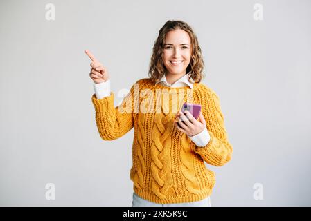 Lächelnde Junge Frau, Die Das Telefon Benutzt Und Zur Seite Zeigt. Ein glückliches, junges Model lächelt, während es ihr Handy benutzt und vor weißem Hintergrund zeigt Stockfoto