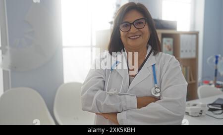 Selbstbewusste hispanische Ärztin mittleren Alters mit kurzen Haaren und Brille steht mit überkreuzten Armen in einem gut beleuchteten Krankenhauszimmer. Stockfoto