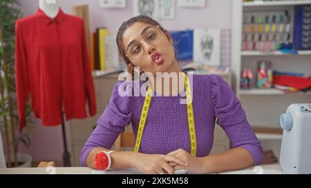 Eine verspielte hispanische Frau in einem lila Pullover macht in einer gut organisierten Schneiderei einen Stupf mit einer roten Bluse auf einer Schaufensterpuppe im Hintergrund. Stockfoto