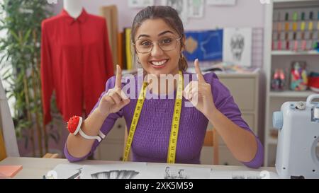 Lächelnde Frau mit Brille, die Zeigefinger in einer bunten Schneiderei mit Schaufensterpuppe und Nähmaschine nach oben bewegt. Stockfoto